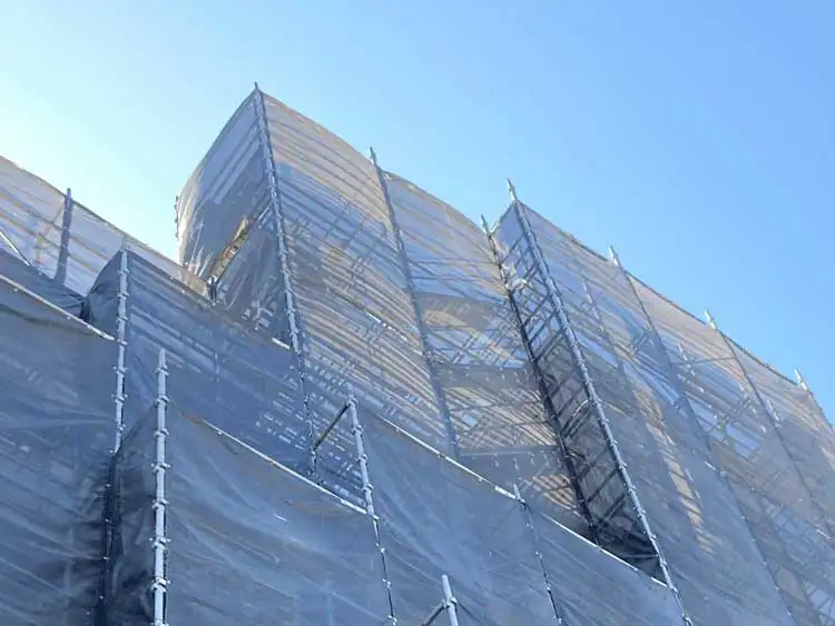 クサビ足場を全面に設置した建設現場の実際の写真。養生シートを施した高層足場の構造が分かる現場風景。
