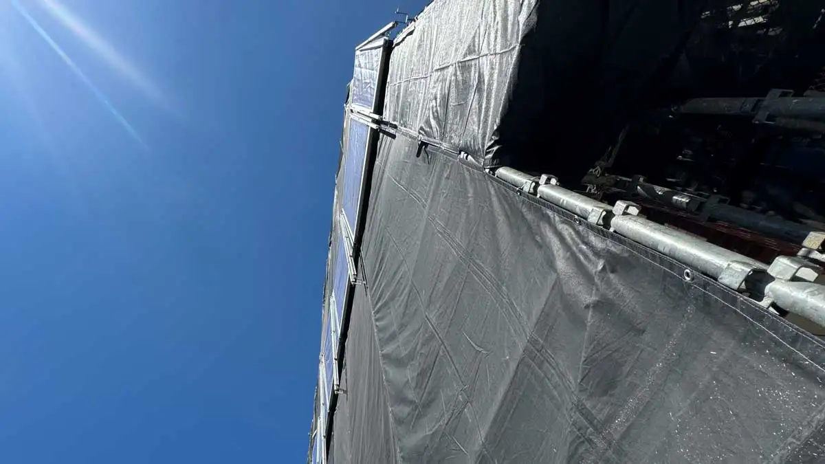 晴天の下、メッシュシートで養生されたくさび式足場を下から見上げた写真。外部足場の安全対策や施工状況を説明する記事用の画像。