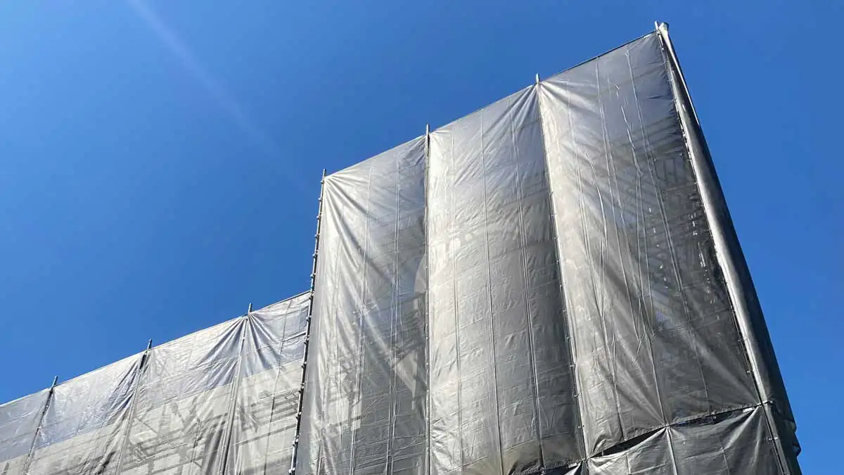 「青空の下で養生シートが張られたクサビ式足場の外部足場全景。住宅外壁工事のための養生状態を示す現場写真。」